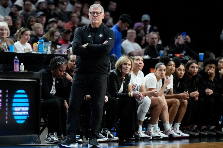 UConn coach Geno Auriemma standing on the sideline during a game with his arms folded.